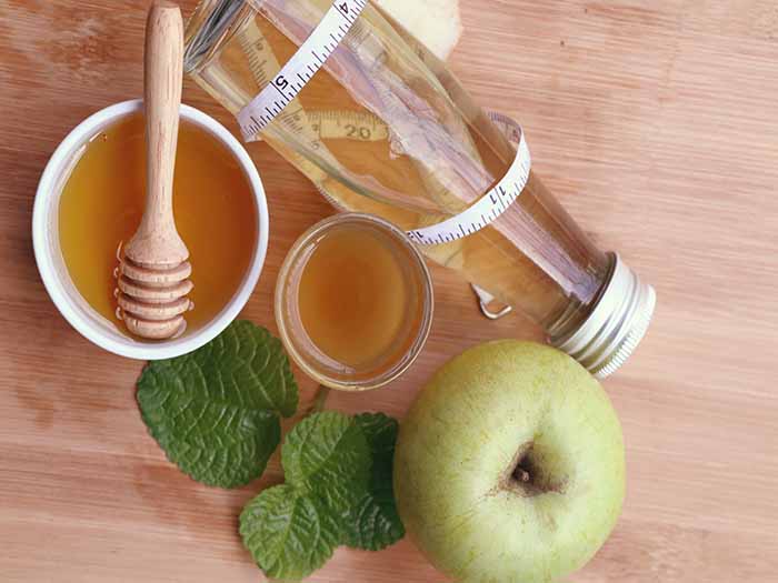 Apple cider vinegar shot in a glass, with an apple, ACV, and honey on a wooden counter