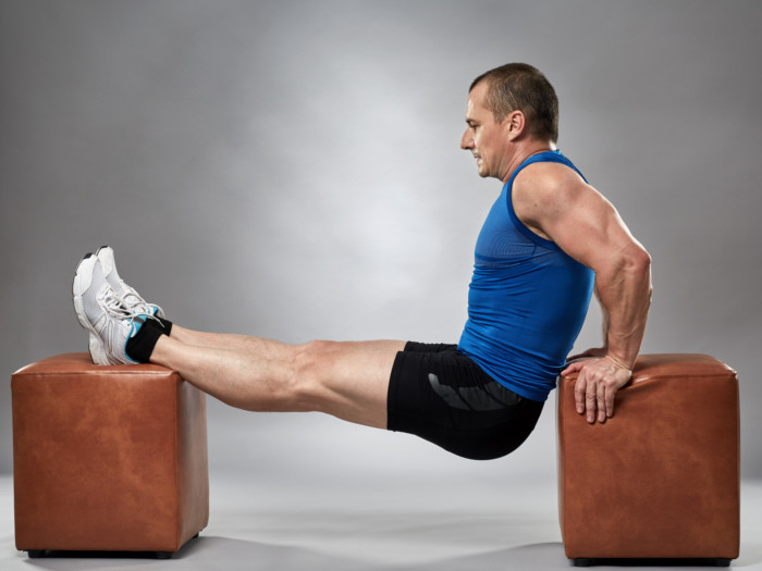 A man in gym clothes performing dips between two brown stools.
