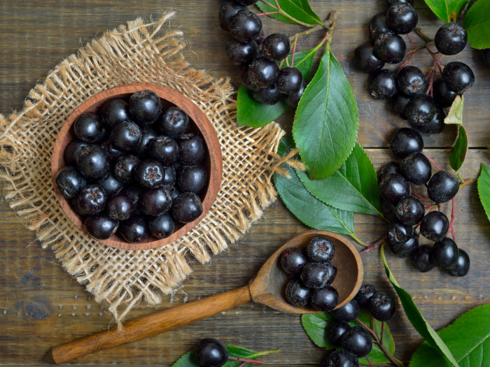 A wooden bowl and spoon of fresh chokeberries with leaves on a wooden table