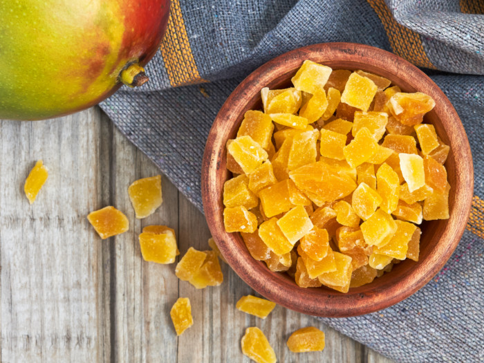 A wooden bowl with sliced dried mango pieces and a whole fresh mango on a wooden table