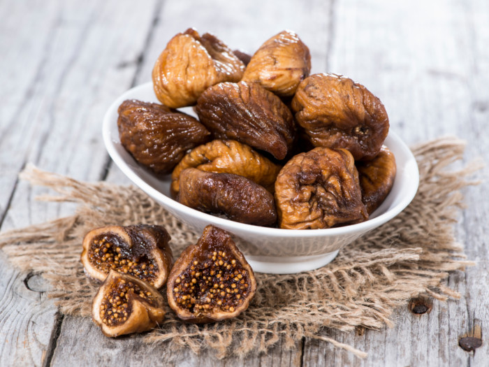 A bowl of dried fig in a white bowl, alongside few more dried figs, placed on a wooden surface.
