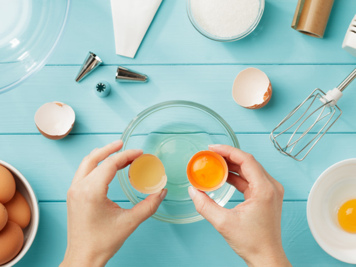 Hand holding eggshells over a bowl, surrounded by a whisk, eggs, piping bag, and top, placed on a light blue wooden surface