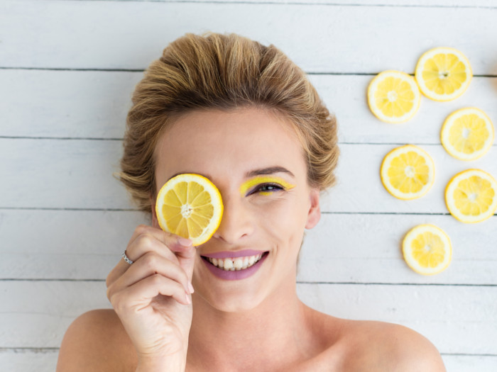 A flat lay pic of a smiling woman covering one of her eyes with a lemon slice