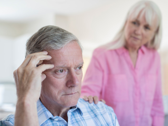 An old man holding his head with a woman at his back