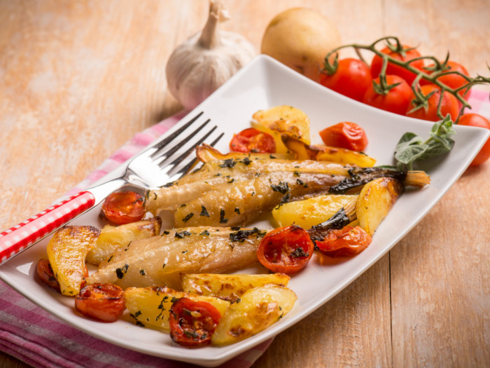 Cooked fish with veggies and potatoes on a plate, served on a wooden surface with tomatoes on the vine, a potato, and garlic.