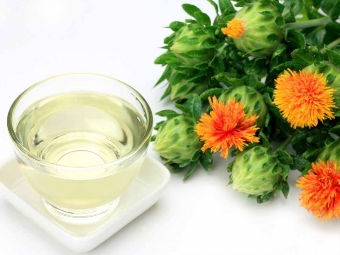 A glass of safflower oil kept next to safflowers against a white background