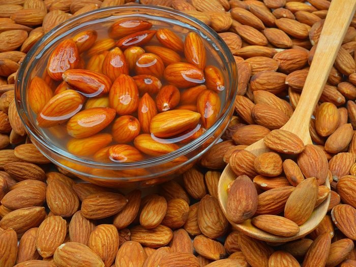 A bowl of soaked almonds with dried almonds on a wooden spoon and background