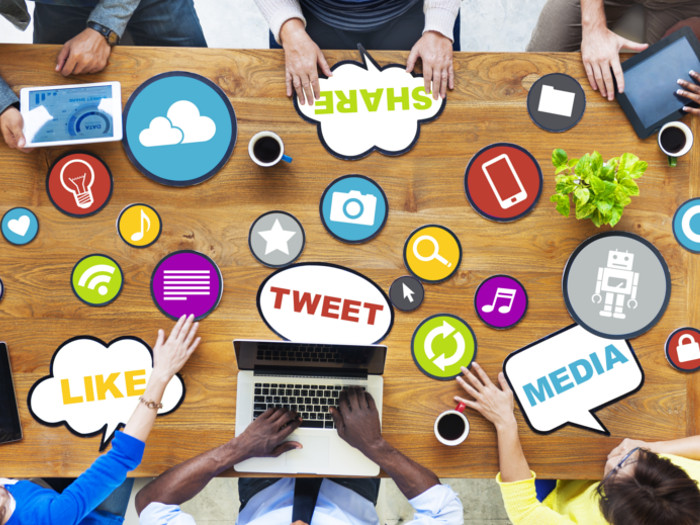 A flat-lay picture of a wooden table covered with logos of different social media platforms and six people working on their laptops