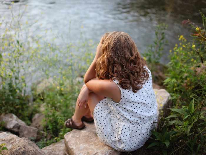 A woman in a polka dot dress with her hand on her forehead, sitting by a stream