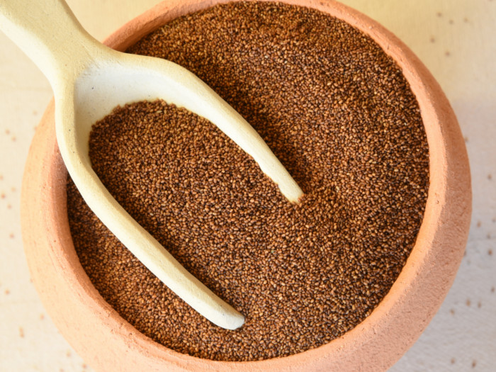 A rustic pottery bowl with a handmade ceramic scoop of teff grains on a white background