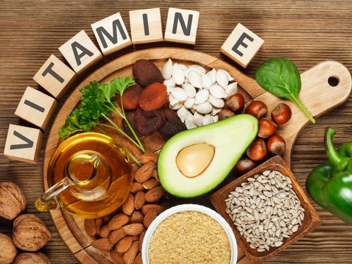 Foods rich in vitamin E (wheat germ oil, dried wheat germ, dried apricots, hazelnuts, almonds, parsley leaves, avocado, walnuts, pumpkin seeds, sunflower seeds, spinach & capsicum) on a wooden table